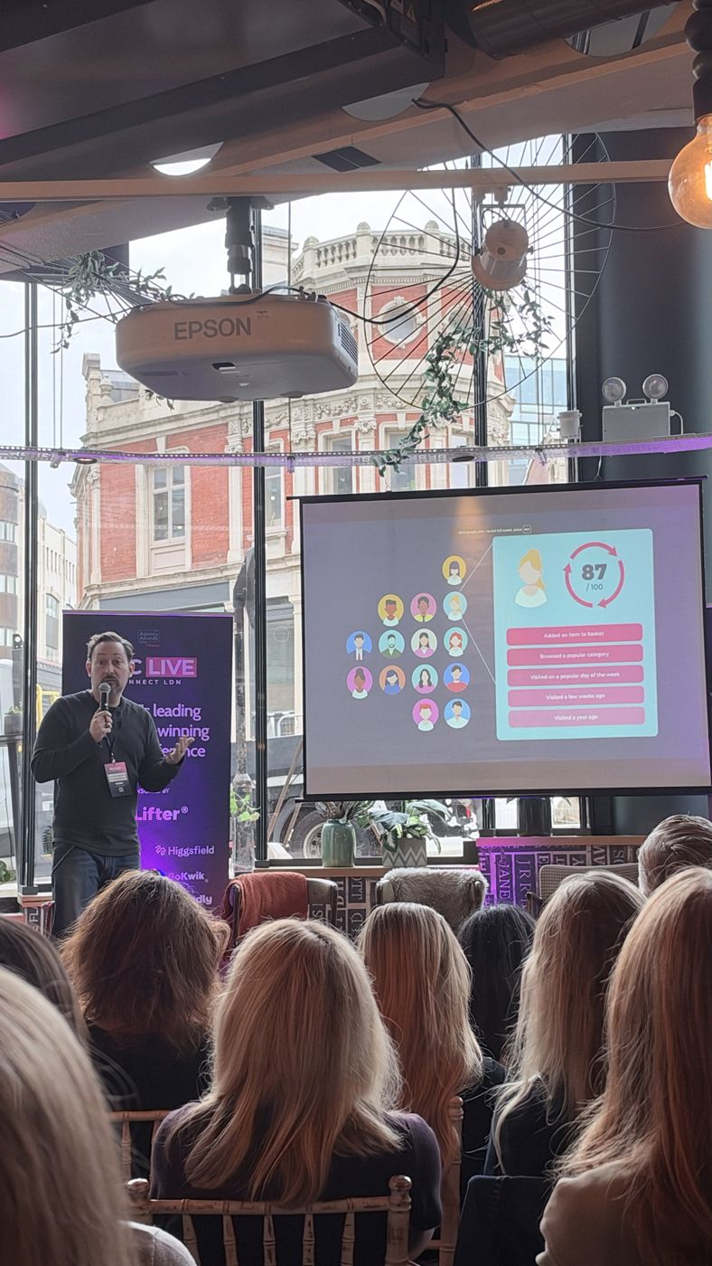 RevLifter speaker on stage beside a slide scoring a shopper 87 out of 100 based on behaviour such as adding to basket and browsing a popular category